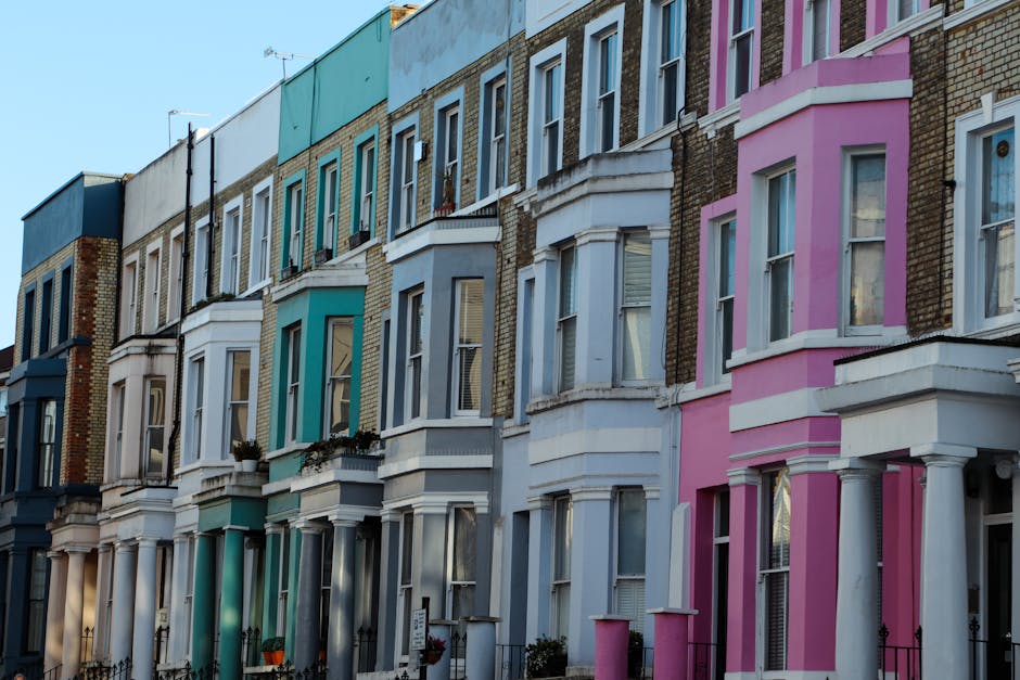 A row of multi-story residential buildings in Notting Hill with a variety of pastel-colored facades including shades of blue, pink, and white. The buildings feature large sash windows, some with flower pots on window sills and small front gardens with wrought iron fences. The surfaces include brickwork and painted exteriors, appearing clean and well-maintained under natural daylight, highlighting the intricate architectural details. This image reflects outdoor residential architecture, with no cleaning tools or surfaces visible, emphasizing the vibrant street aesthetic associated with the area. Carpet Cleaning Notting Hill specializes in surface cleaning and deep cleaning services to maintain the hygiene and appearance of similar residential exteriors.
