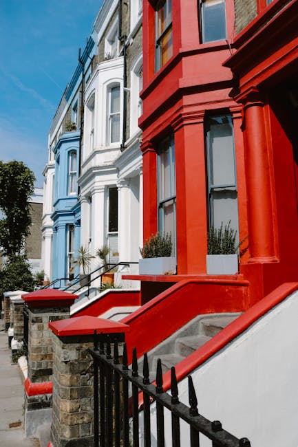 A row of Victorian terraced houses in Notting Hill with brightly painted facades, including pastel blue and vibrant red. The houses feature large sash windows and decorative architectural details. Each property has a small front garden or steps leading to the entrance, with black wrought iron railings and stone steps. The street is lined with trees, and the sky is clear with natural daylight illuminating the scene. The image emphasizes the exterior surfaces such as painted brickwork and stone steps, showcasing cleanliness and well-maintained appearances, consistent with professional domestic and commercial surface cleaning standards promoted by Carpet Cleaning Notting Hill.