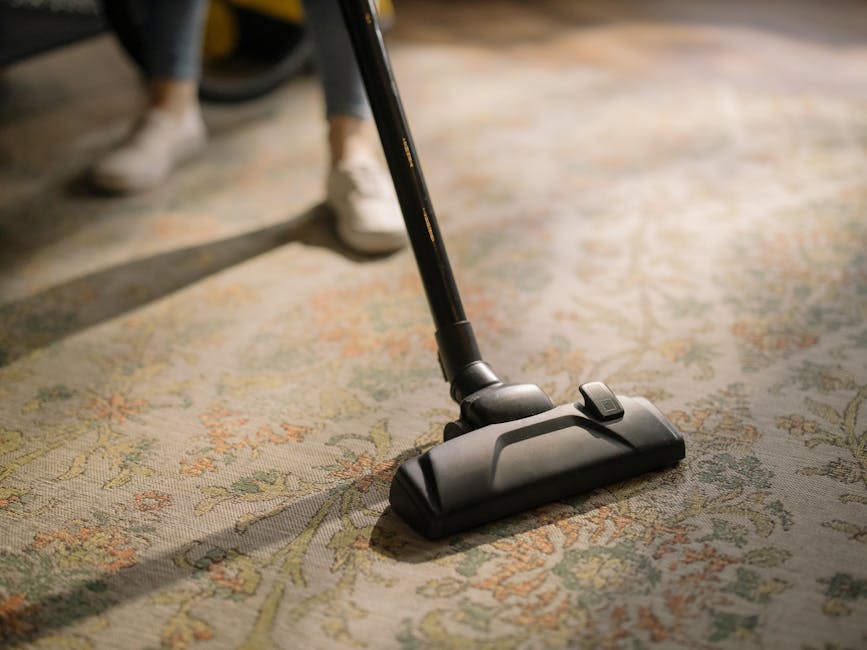 Close-up of a vacuum cleaner attachment in use on a patterned carpet within a home setting. The vacuum head is black with a textured surface and is connected to a long black hose or extension wand. The carpet features intricate floral motifs in shades of beige, green, and light orange, appearing clean and well-maintained. In the background, a person's legs and white sneakers are partially visible, suggesting active carpet cleaning or preparation work, aligned with estate agent carpet prep for Holland Park listings. The room is illuminated with natural light, highlighting the surface cleanliness. This image illustrates professional surface cleaning processes carried out by Carpet Cleaning Notting Hill as part of domestic cleaning services.