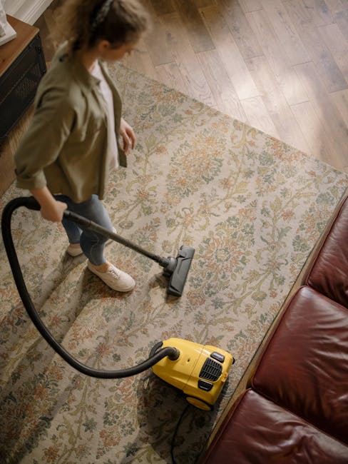 A woman using a yellow vacuum cleaner to perform surface cleaning and deep cleaning on a patterned area rug in a living room. The rug features floral motifs in shades of beige, green, and orange, laid on a wooden floor with a polished, light brown finish. She is dressed casually in a beige jacket, white shirt, and jeans, focusing on the task with attention to hygiene and maintenance. Nearby, there is a red leather sofa, and the room is well-lit with natural light, emphasizing cleanliness and meticulous care typical of domestic cleaning services by Carpet Cleaning Notting Hill.