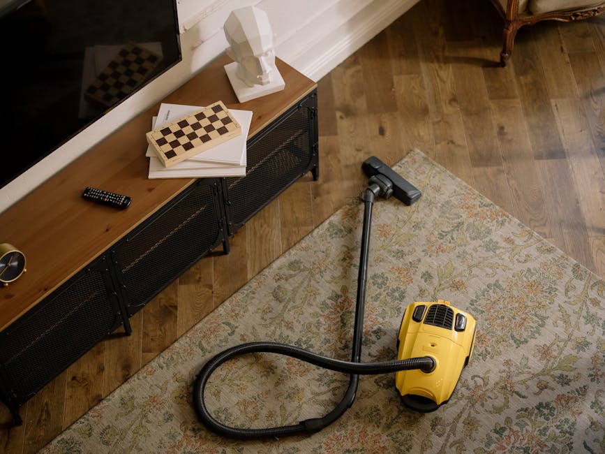 Close-up of a vacuum cleaner attachment in use on a patterned carpet within a home setting. The vacuum head is black with a textured surface and is connected to a long black hose or extension wand. The carpet features intricate floral motifs in shades of beige, green, and light orange, appearing clean and well-maintained. In the background, a person's legs and white sneakers are partially visible, suggesting active carpet cleaning or preparation work, aligned with estate agent carpet prep for Holland Park listings. The room is illuminated with natural light, highlighting the surface cleanliness. This image illustrates professional surface cleaning processes carried out by Carpet Cleaning Notting Hill as part of domestic cleaning services.
