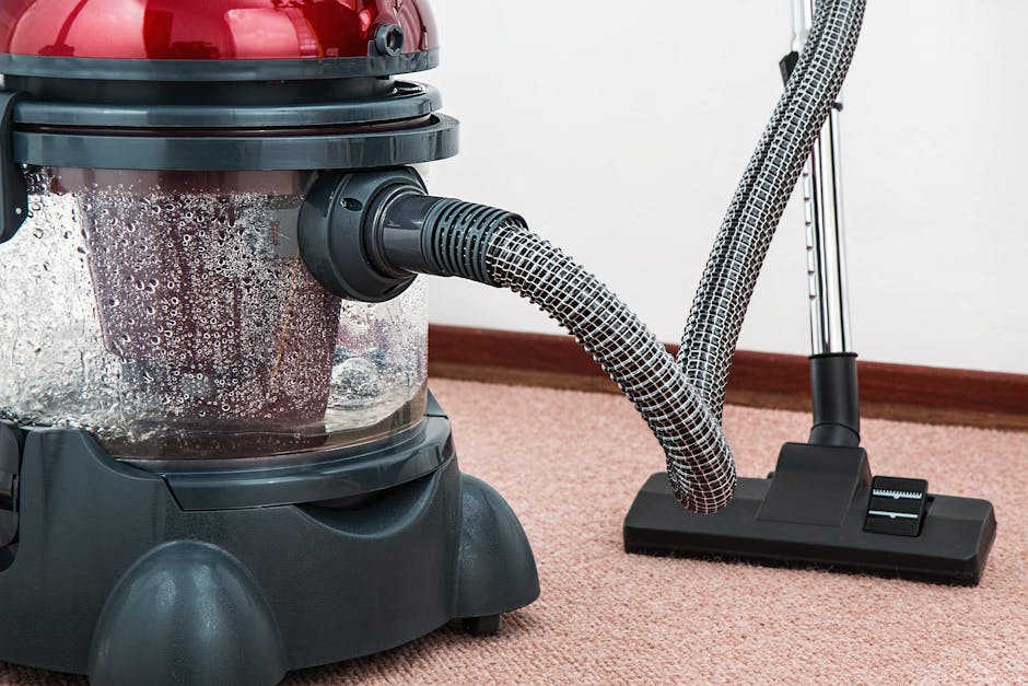 A person holding a yellow portable air mover or blower in a room with a worn, patterned carpet. The carpet features a floral design with muted tones of beige, green, and orange. The individual’s han