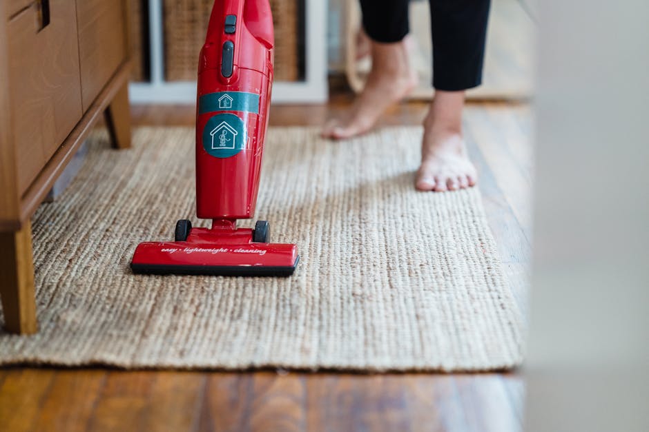 Close-up of a person using a red handheld vacuum cleaner to clean a beige textured area rug in a residential setting, with wooden flooring and furniture visible. The person is barefoot and wearing black pants, emphasizing domestic cleaning activities. The vacuum cleaner bears the logo and name of Carpet Cleaning Notting Hill, indicating a professional cleaning service. The scene is well-lit, highlighting the cleanliness of the carpet and surrounding surfaces, which include polished wooden floors and wooden furniture. This image reflects surface cleaning and maintenance related to hygiene and domestic cleaning, consistent with the guidelines on carpet disposal and cleaning rules for the Kensington & Chelsea area, as outlined by the [DOMAIN] website.