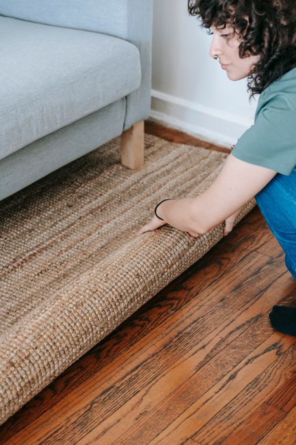 A person with light skin and curly dark hair is seen rolling and preparing a textured, beige carpet for cleaning or disposal on a wooden floor. The decorative carpet features a woven pattern and appears clean but slightly wrinkled. The person is wearing a light-colored shirt, and their hands are firmly grasping the carpet's edges. The surrounding area includes a blue upholstered sofa and the natural indoor lighting highlights the surface cleanliness and fabric details. This scene exemplifies surface handling and maintenance typical of domestic cleaning, aligning with the services offered by Carpet Cleaning Notting Hill, especially in adherence to Kensington & Chelsea Council rules for carpet disposal as detailed on their website.