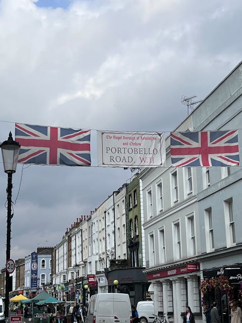 A busy street scene in Notting Hill, London, featuring a row of white and light-colored terraced buildings with various retail shops on the ground floor. The street is active with pedestrians, some en