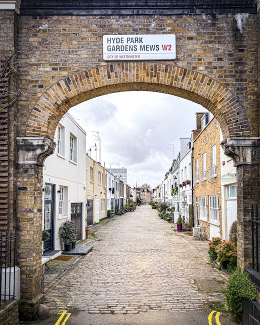 A cobblestone street view through an arched brick gateway with a sign reading 'HYDE PARK GARDENS MEWS W2' at the top, leading into a residential area with white and light-colored terraced houses, some with doors and small potted plants outside. The street appears clean and well-maintained with no visible litter or stains, suggesting regular surface cleaning and upkeep, as part of typical property and street maintenance in this London neighborhood. The sky is partly cloudy, providing natural light that highlights the fresh, tidy appearance of the facades and pavement with no visible dirt or debris, in line with the cleaning and sanitisation standards promoted by Carpet Cleaning Notting Hill in their domestic cleaning and surface care advice.