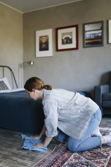 A woman with brown hair tied back, dressed in a light blue and white striped long-sleeve shirt and jeans, kneeling on a patterned rug in a living room, carefully cleaning a dark blue upholstered bed frame with a cloth. The room features a wooden floor with a decorative rug, beige walls decorated with framed artwork, a gray armchair, and a bedside table, all illuminated by natural light. This scene illustrates domestic surface cleaning and maintenance, highlighting the importance of regular hygiene and deep cleaning by professional services such as Carpet Cleaning Notting Hill, as recommended in advice on stain care for Pembridge Villas flats and mews.