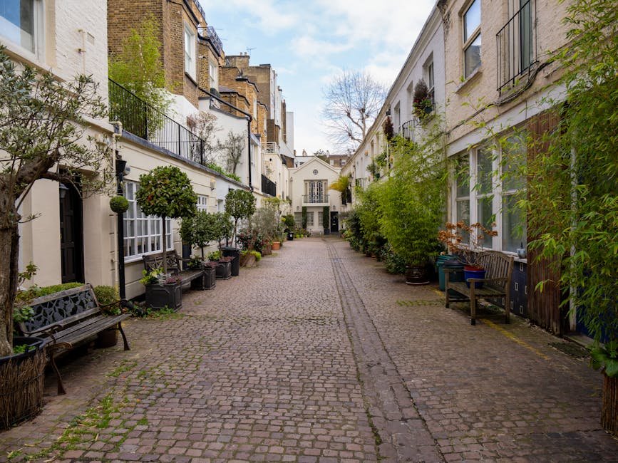 A cobblestone street view through an arched brick gateway with a sign reading 'HYDE PARK GARDENS MEWS W2' at the top, leading into a residential area with white and light-colored terraced houses, some with doors and small potted plants outside. The street appears clean and well-maintained with no visible litter or stains, suggesting regular surface cleaning and upkeep, as part of typical property and street maintenance in this London neighborhood. The sky is partly cloudy, providing natural light that highlights the fresh, tidy appearance of the facades and pavement with no visible dirt or debris, in line with the cleaning and sanitisation standards promoted by Carpet Cleaning Notting Hill in their domestic cleaning and surface care advice.