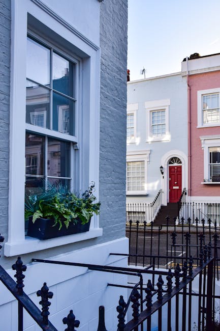 A close-up view of a residential window with white framing and a flower box filled with green plants positioned on the windowsill. The exterior wall is painted light blue, and the window has clear gla